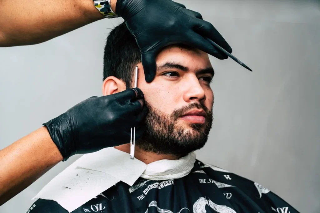 A barber wearing black disposable gloves uses a straight razor to carefully line up a man's hair and beard in a barbershop